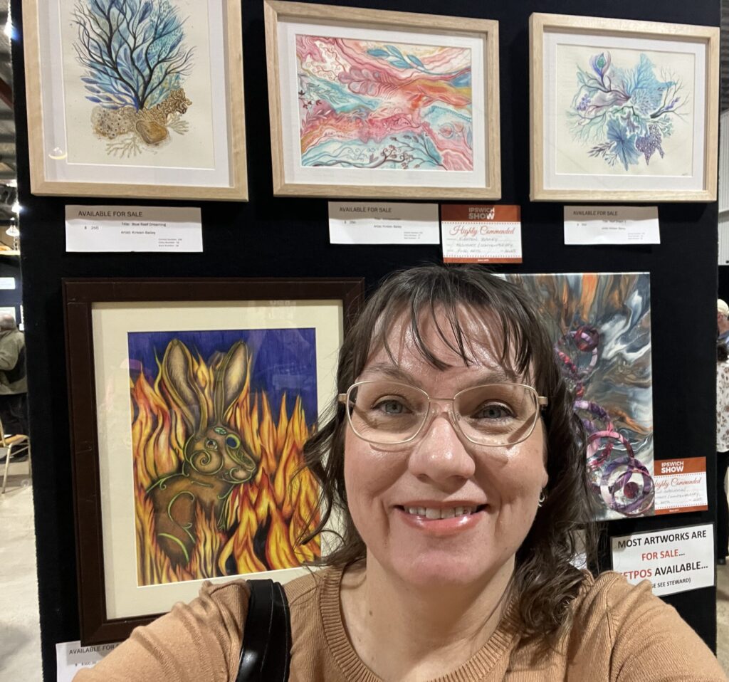 Artist Kirsten Bailey standing in front of 3 of her paintings hanging up at the Ipswich Show